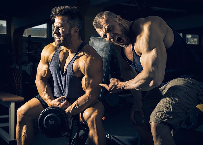 Two muscular men at the gym, one lifting weights while the other yells, highlighting tension and confrontation.