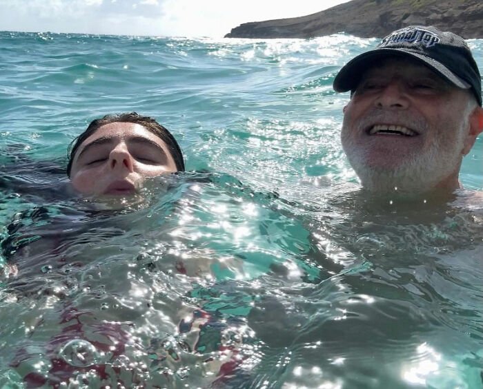 Rob Reiner's daughter and father swimming in ocean waves under clear sky near rocky coastline.
