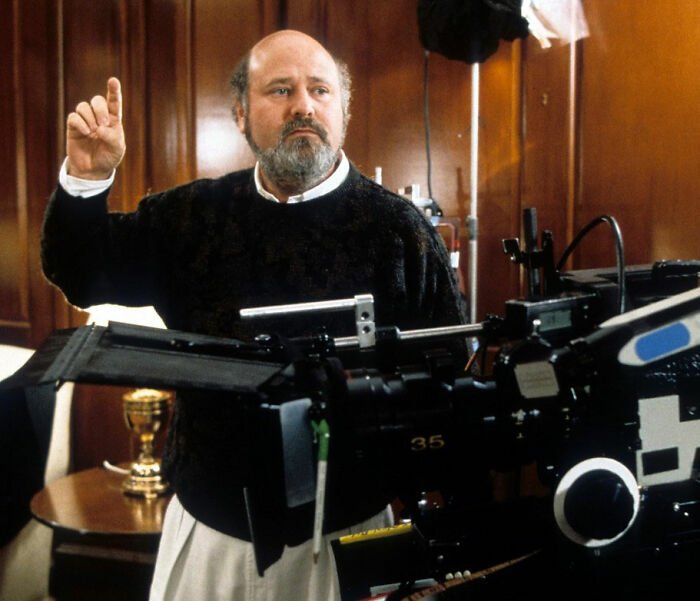 Rob Reiner directing on set, wearing a dark sweater, behind camera equipment with a wood paneled background.