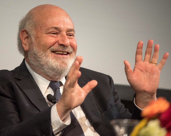 Rob Reiner speaking at an event, smiling and gesturing with hands while wearing a suit and tie.