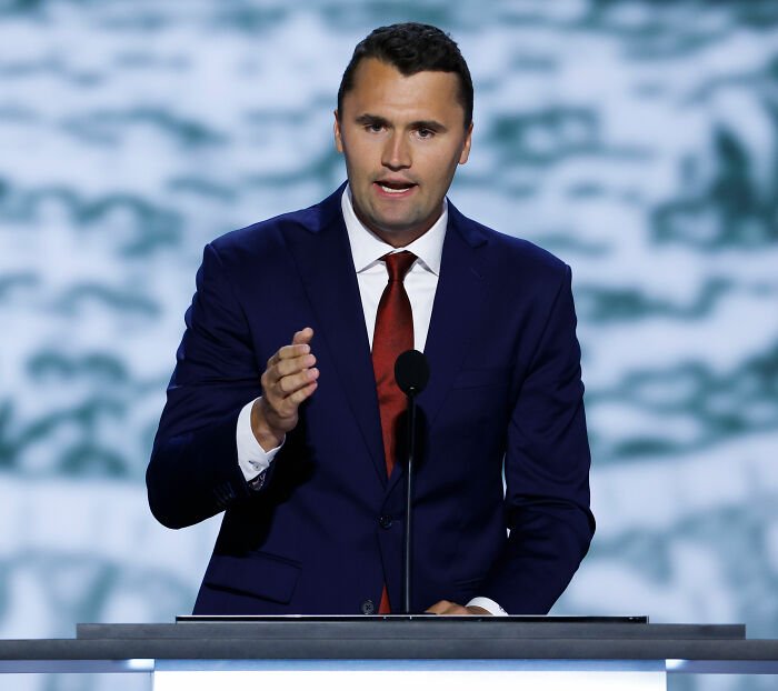 Man in blue suit and red tie speaking at a podium, related to MAGA hypocrisy debate and political commentary.