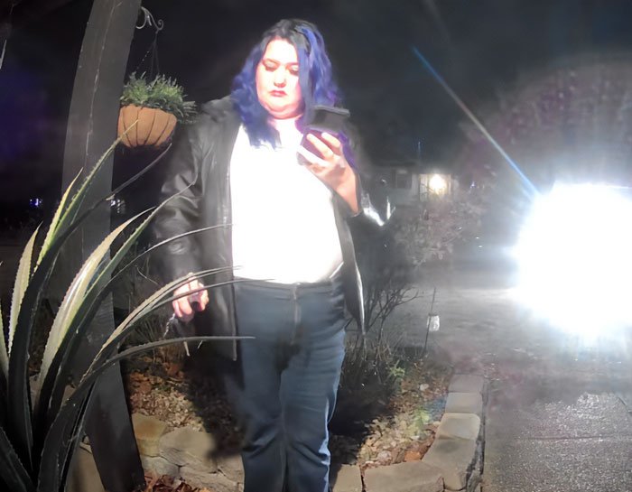 Woman with blue hair wearing a black jacket seen on a porch at night near plants and bright light source.