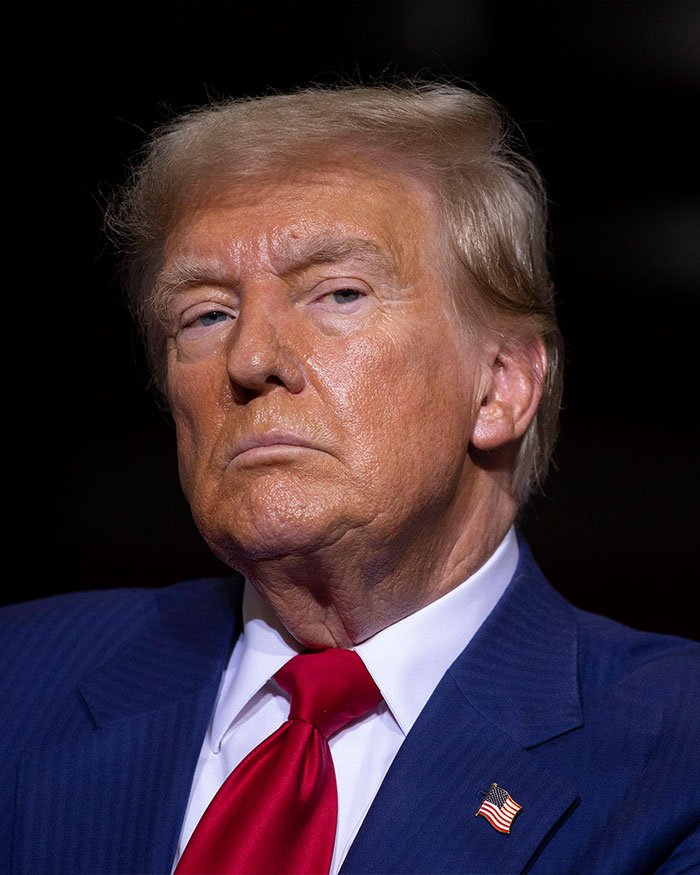 Donald Trump in a blue suit and red tie, facing forward with a serious expression during a public appearance.