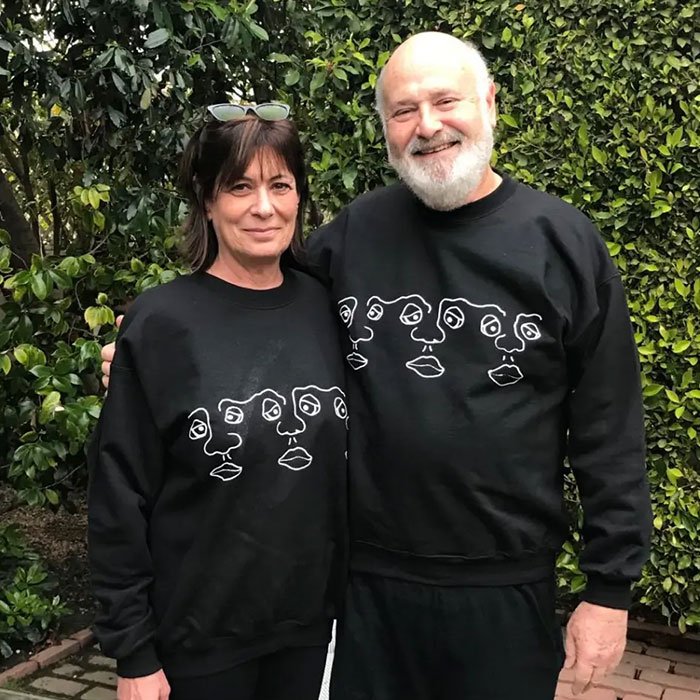 Rob Reiner and wife standing outdoors wearing matching black sweatshirts with abstract face designs.