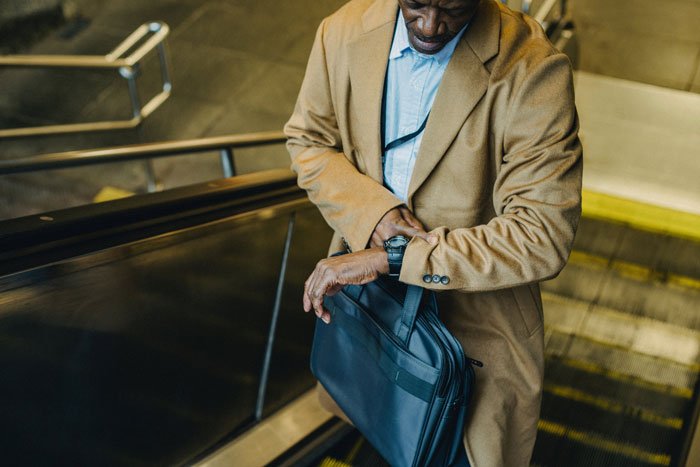 Man with two jobs checking watch on escalator, carrying briefcase, symbolizing busy work life and family challenges.