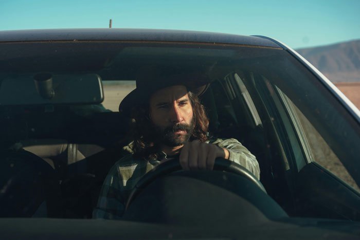 Man with two jobs driving and looking serious inside car, reflecting tension around jobless wife and son not ready for daycare.