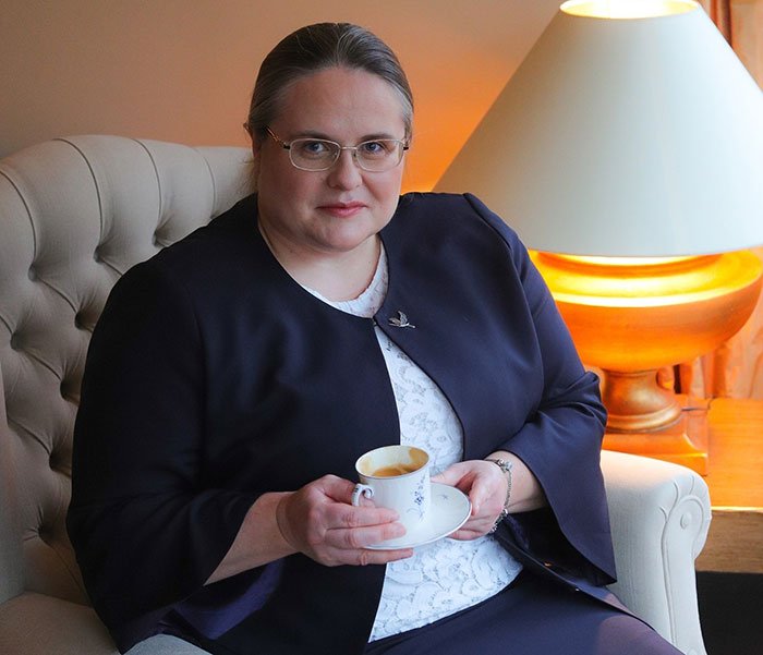 Woman in glasses holding a cup, seated indoors near a lamp, related to Lithuania lawmaker’s cat consent news.