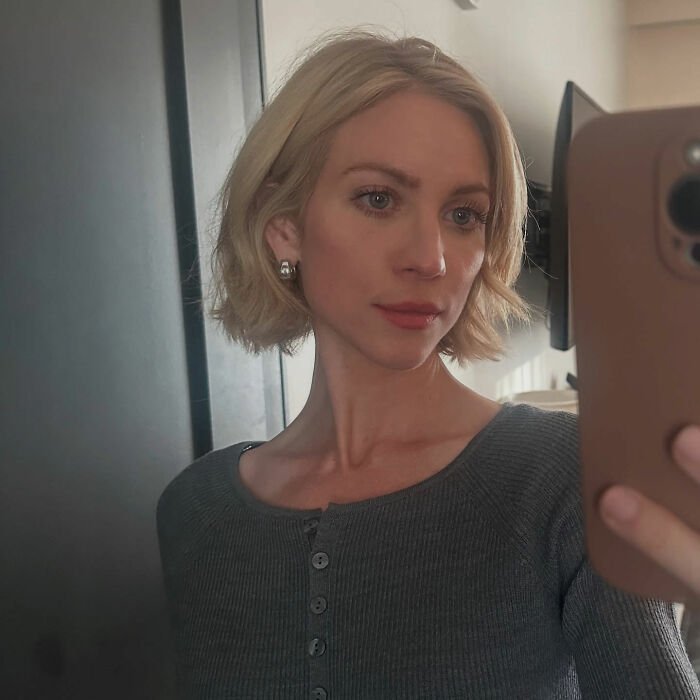 Britanny Snow taking a selfie indoors with short blonde hair, wearing earrings and a grey buttoned top, addressing plastic surgery rumors.