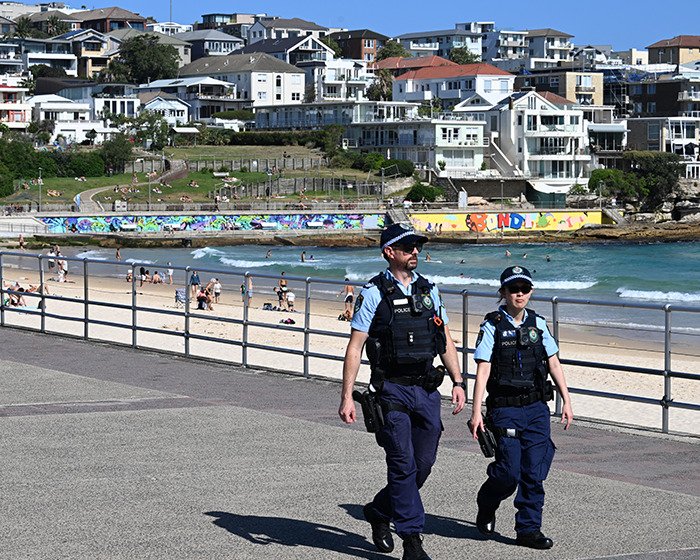 Two police officers walking near Bondi Beach with beachgoers and residential buildings in the background on a sunny day