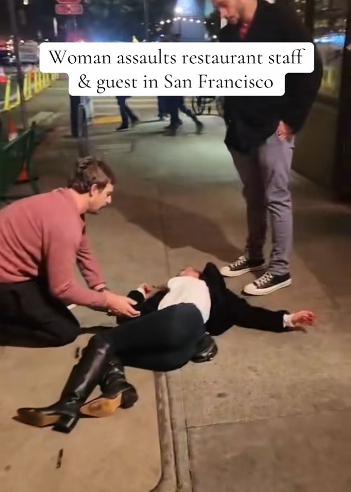 Woman lying on sidewalk after drunken rampage at upscale restaurant, helped by bystanders in urban night scene.
