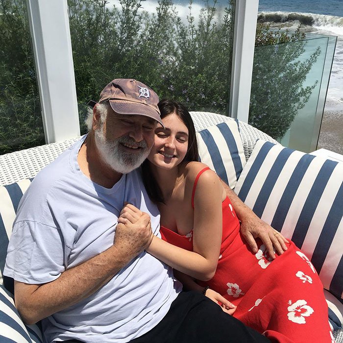 Rob Reiner&rsquo;s children Jake and Romy embracing outdoors on striped cushions near greenery and ocean waves.