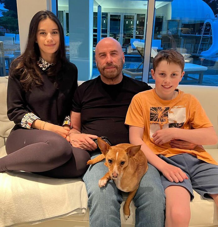 Riley Keough, John Travolta, and son Ben sitting on a couch with a small dog indoors, casual family moment