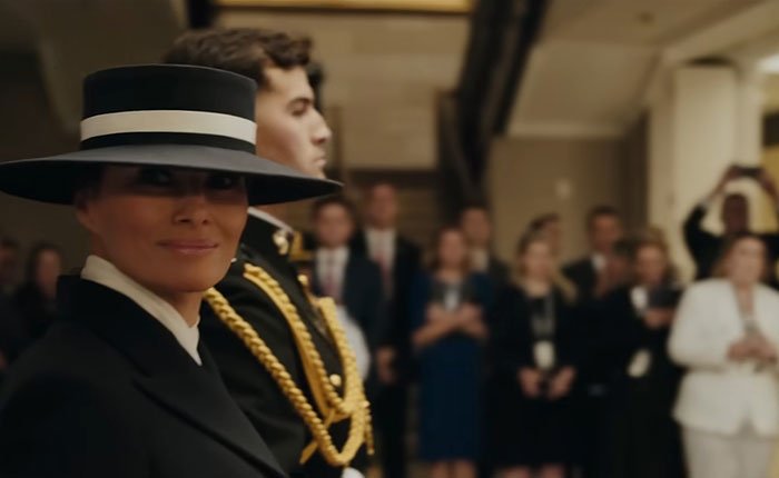 Melania Trump wearing a black hat and coat at a formal event with people in the background.