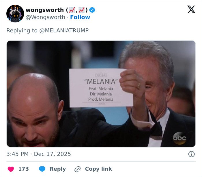 Two men in tuxedos holding up a card humorously listing Melania as director, producer, and feature of a documentary.