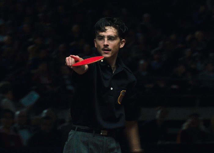 Timothée Chalamet playing ping pong wearing glasses and a black shirt in a dimly lit setting.