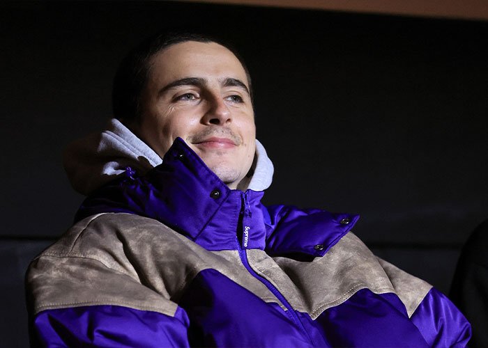Man wearing a purple and beige jacket, smiling slightly, in a dark indoor setting related to Timothée Chalamet arrogance controversy.