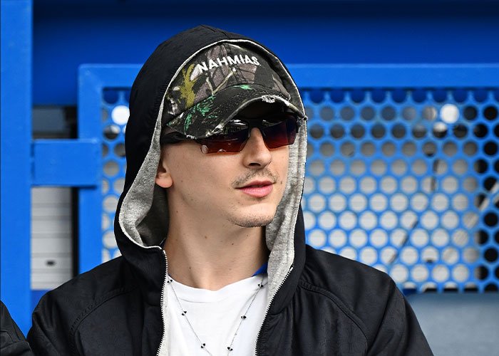 Timothée Chalamet wearing sunglasses and a cap, facing sideways with a casual black hoodie in an outdoor setting.