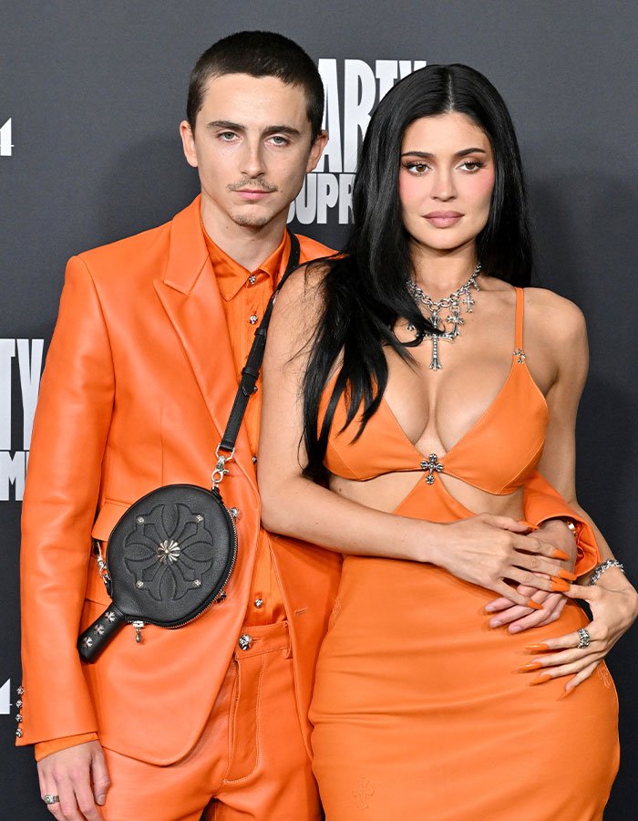 Timothée Chalamet wearing orange suit posing with woman in orange dress at an event, highlighting arrogance controversy.