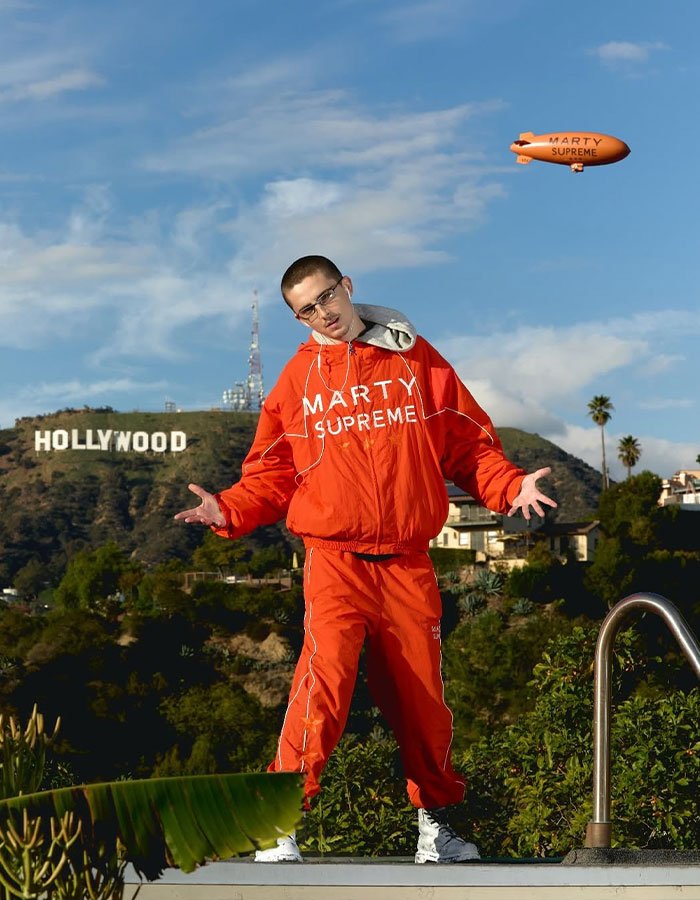 Man in red tracksuit posing near Hollywood sign under blue sky during daytime, reflecting Timothée Chalamet arrogance controversy.