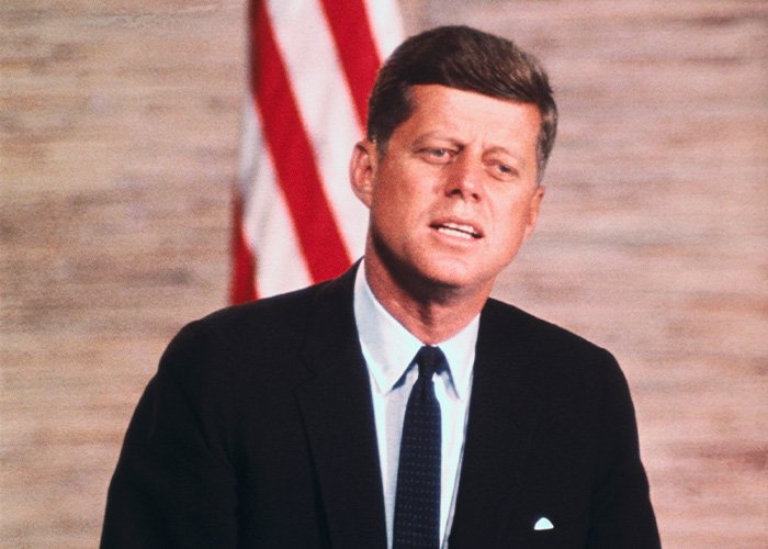 Man in a dark suit speaking in front of a blurred American flag, related to Kennedy Center renaming controversy online.