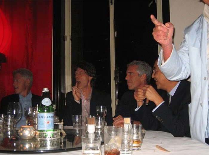 Jeffrey Epstein and others seated at a table with drinks as chilling toddler's foot is visible beside Epstein in photo.