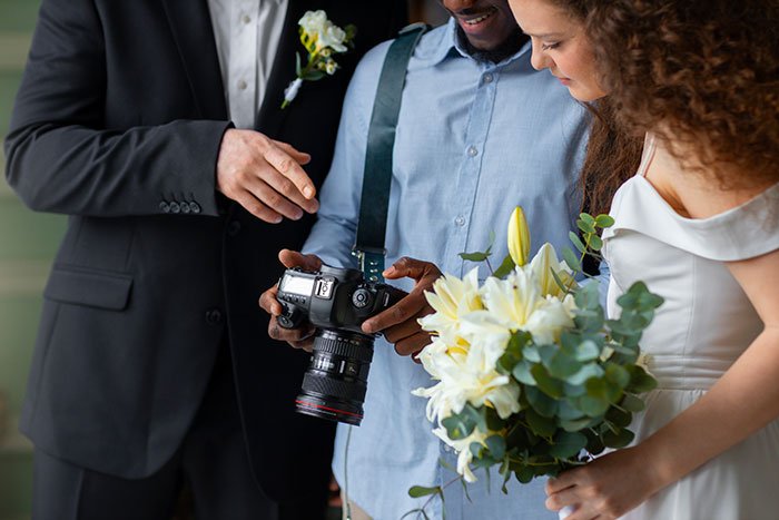 Bride and groom reviewing wedding photos with photographer, bride holding white lily bouquet, wedding day preparations.