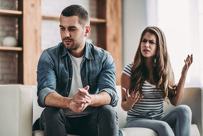 Woman looks upset while fiancé sits silently on couch worried about their wedding photos and appearance.