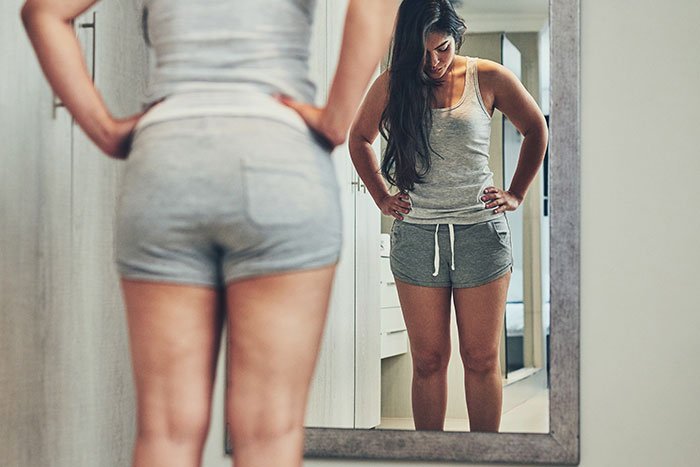 Woman in gray tank top and shorts looking worriedly at her reflection, concerned about appearance before wedding photos.