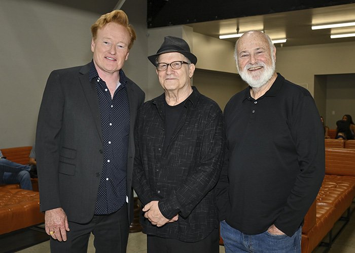 Conan O’Brien with two men at an event, posing for a photo in a room with leather seating and industrial lighting.