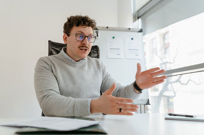 Man in gray sweater explaining open marriage issues with expressive hand gestures in a bright office setting.