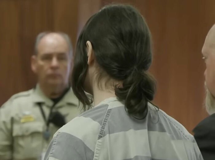 Trans Muslim woman in court wearing prison uniform during sentencing hearing with officers and legal counsel present.