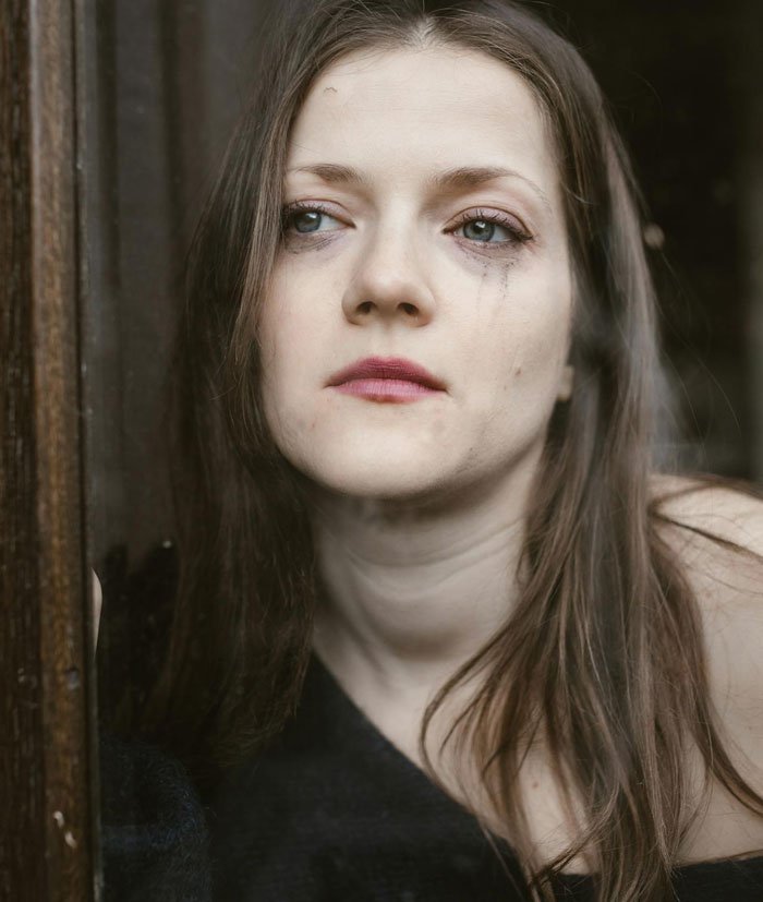 Woman with smudged makeup looking out a window, reflecting on uncertain affair partner's baby and complex family ties.