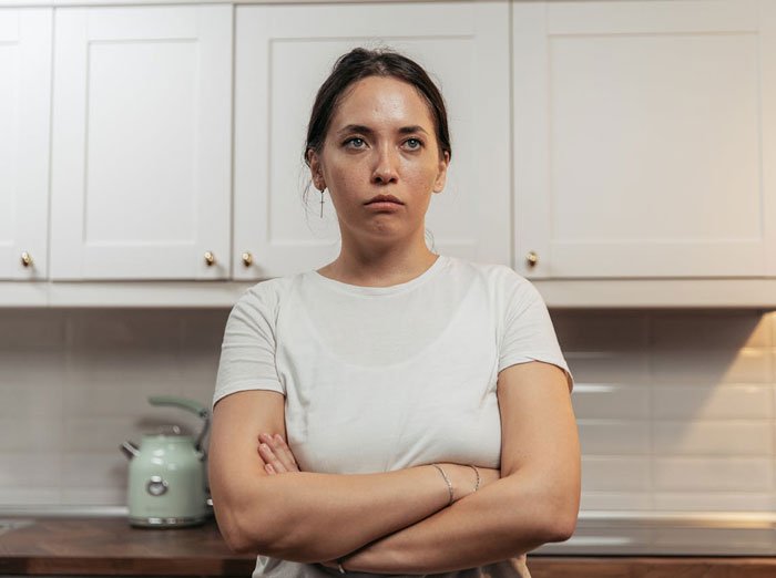 Young woman with crossed arms and a serious expression in a kitchen, reflecting uncertainty about family relationships.