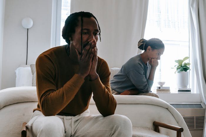 Man sitting on couch looking unsure while woman in background appears upset, reflecting affair partner baby paternity doubts.