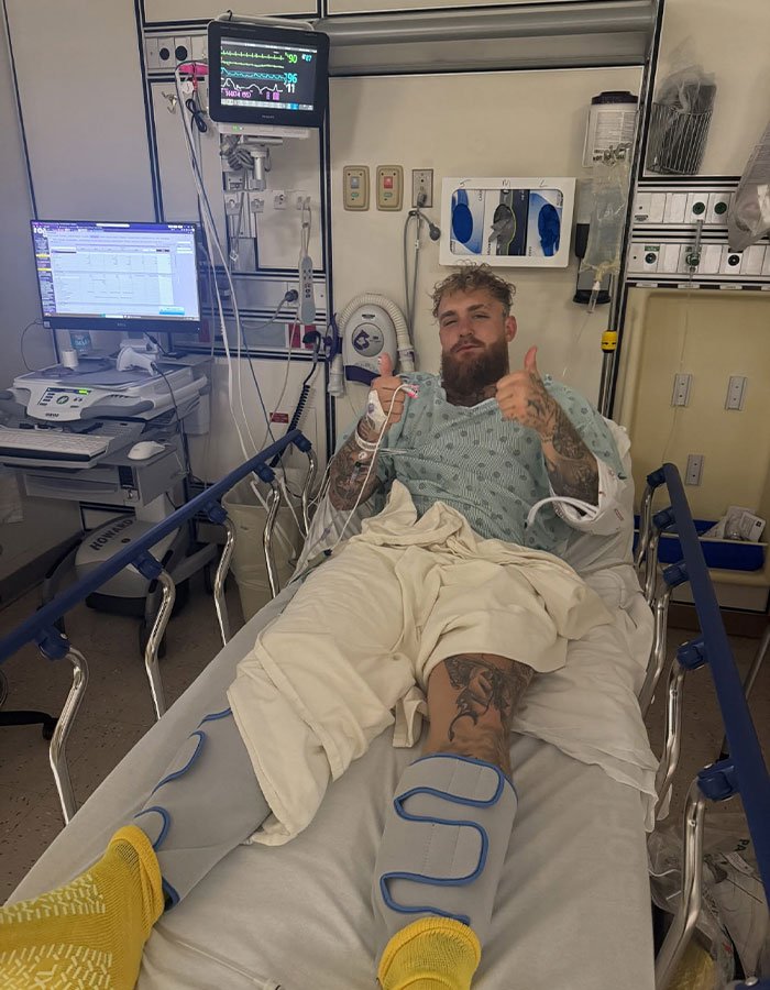 Man in hospital bed with leg braces giving thumbs up, highlighting Jake Paul's broken jaw trending on the internet.
