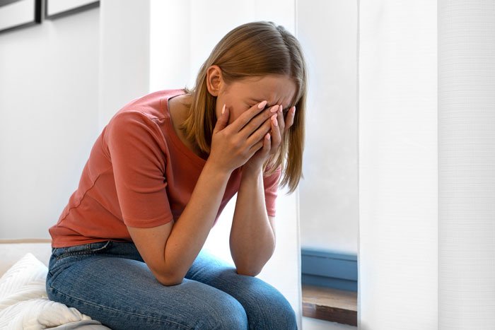 Woman sitting on bed with face in hands, appearing upset, in a bright room near a window during daytime.