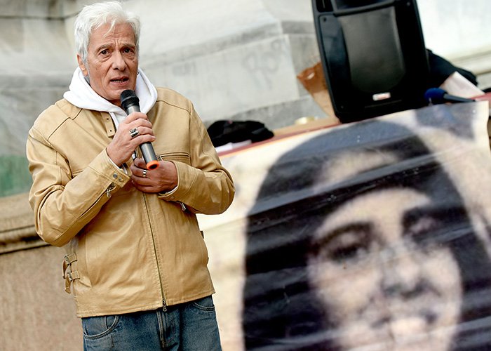 Man in tan jacket speaking into microphone next to large image of the Vatican Girl from decades-old cold case.