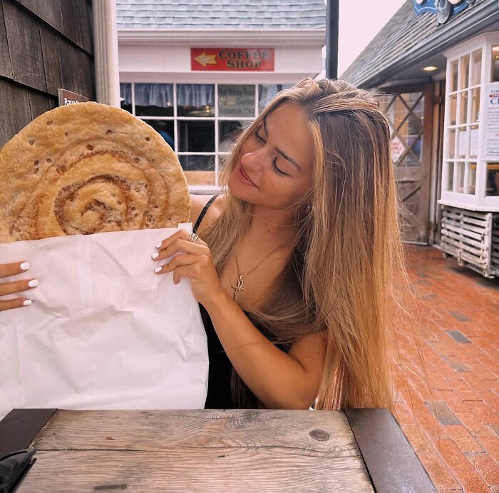 21-year-old pizza maker sitting outdoors, holding large pizza crust wrapped in paper, smiling and looking at it.