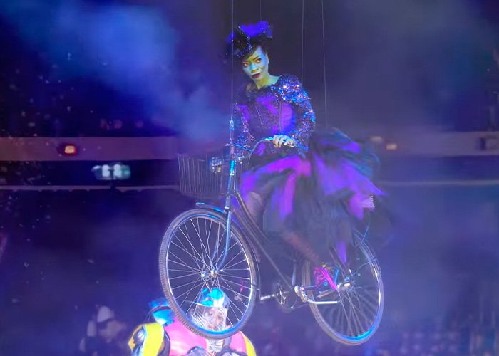 Performer in colorful costume on a suspended bicycle during Wizard of Oz performance amid audience reaction.