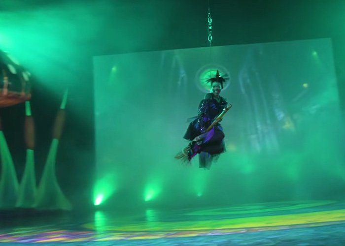 Actor dressed as a witch flying on a broomstick during Wizard Of Oz performance with dramatic green stage lighting and fog effects.