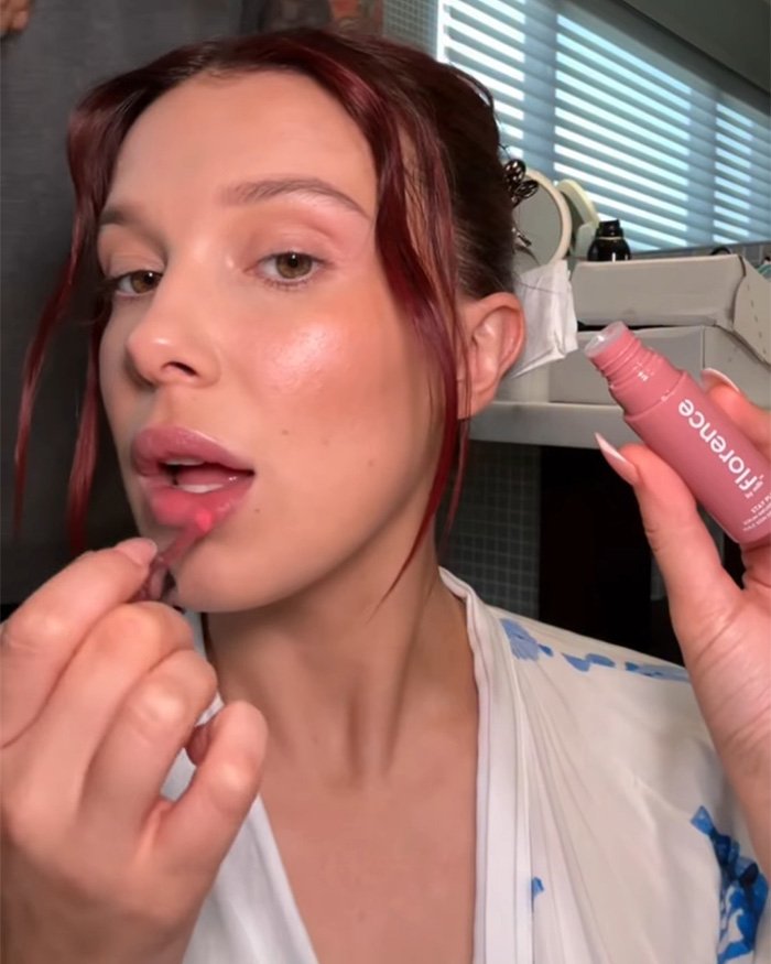 Millie Bobby Brown applying lip gloss in a close-up shot as fans zoom in on her Stranger Things appearance.