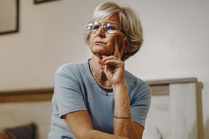 Older woman with glasses thoughtfully resting her face on her hand, reflecting on wedding dress controversy with bride and son.