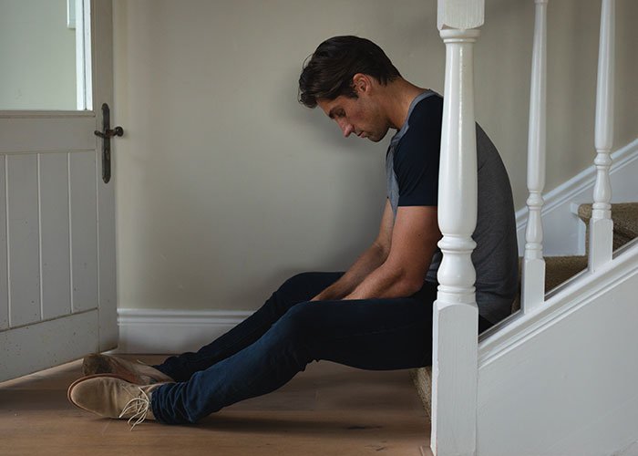 Man sitting on floor near stairs, looking down sadly, representing husband’s ruined reputation after wife leaves him.