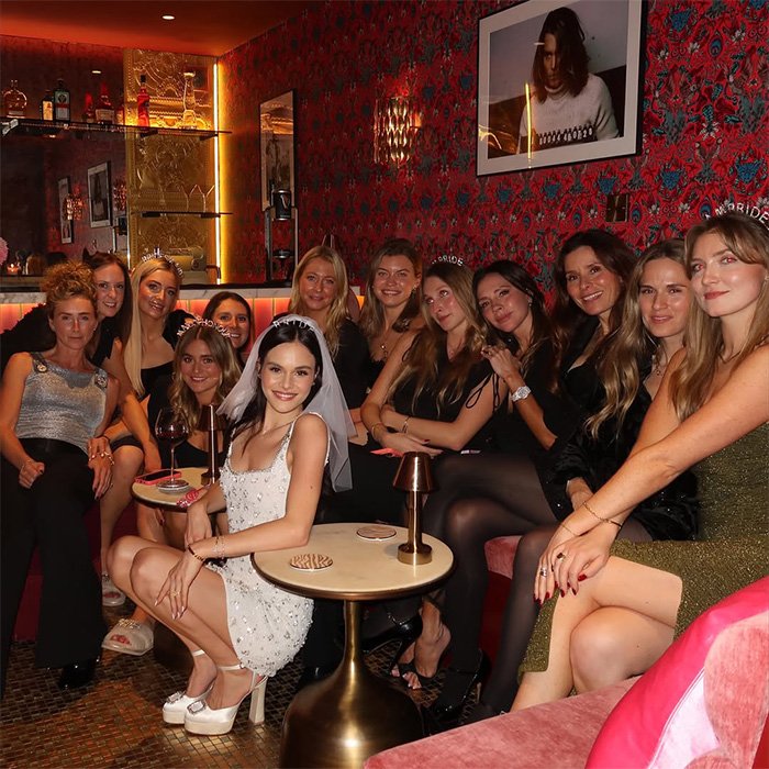 Group of women celebrating indoors with the bride in a satin bed sheet-inspired dress, highlighting wedding buzz and fashion.