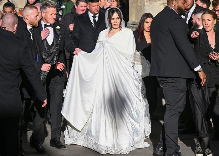 Bride in a white satin bed sheet style wedding dress surrounded by guests at an outdoor event.