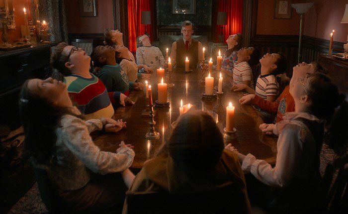 Group of children and a man sitting around a candlelit table in a dark room, Stranger Things 5 scene with eerie atmosphere.