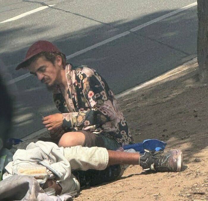 A man in a floral shirt and red cap sitting on the ground next to a person lying down, related to Tylor Chase hospitalization.