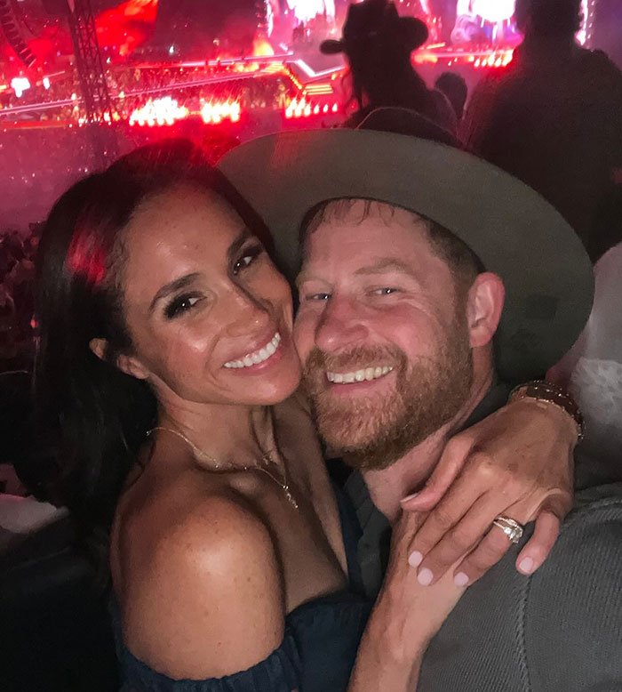 Prince Harry and Meghan Markle smiling together at a crowded event with vibrant red lighting in the background.