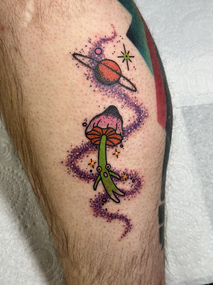 Colorful psychedelic tattoo of a mushroom and planet with stars surrounded by a swirling purple cosmic trail on skin.