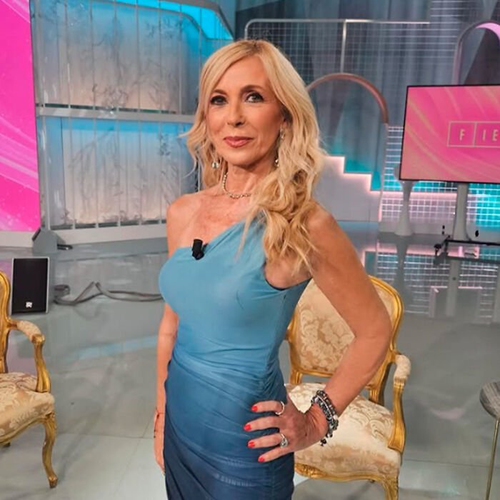 Woman in a blue dress posing indoors with blurred chairs and screens, related to Kate Middleton’s facial scar rumors.